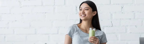 Jeune femme asiatique avec verre de smoothie frais souriant tout en regardant loin, bannière — Photo de stock