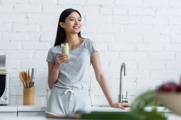 Felice donna asiatica in possesso di un bicchiere di frullato fresco mentre in piedi in cucina — Foto stock