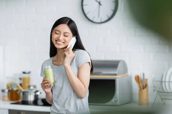 Smiling asian woman with glass of fresh smoothie talking on cellphone in kitchen — Stock Photo