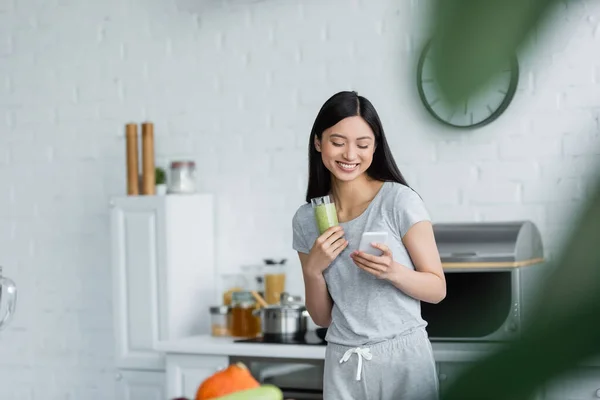 Cheerful asian woman with glass of smoothie messaging on cellphone on blurred foreground — Stock Photo