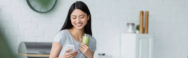 Smiling asian woman with glass of delicious smoothie using smartphone in kitchen, banner — Stock Photo