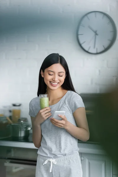 Feliz asiático mujer usando móvil teléfono mientras celebración vidrio de sabroso smoothie - foto de stock