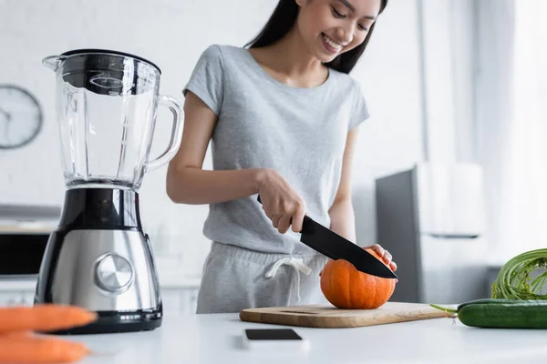 Sorrindo asiática mulher cortando abóbora perto de liquidificador elétrico, smartphone e legumes frescos — Fotografia de Stock