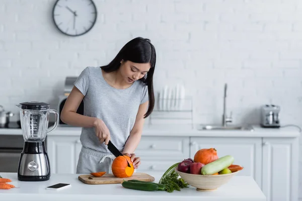 Young asian woman cutting pumpkin near vegetables, electric blender and smartphone — Stock Photo