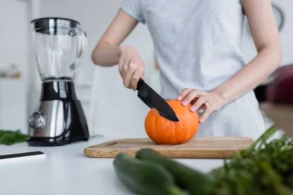 Vue recadrée de femme coupant citrouille mûre près secoueur électrique et concombres flous — Photo de stock