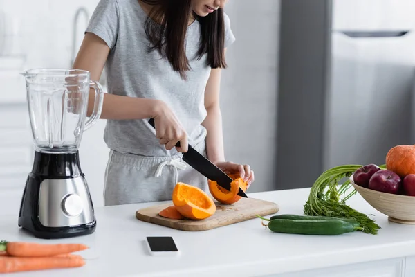 Partial view of woman cutting ripe pumpkin near smartphone with blank screen and shaker — Stock Photo