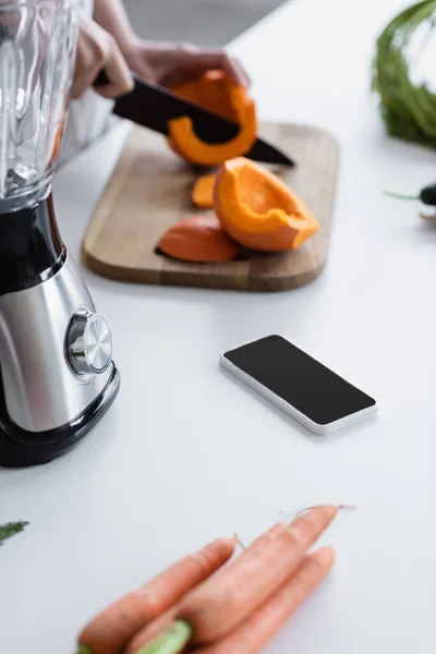 Vista parcial de la mujer borrosa cortando calabaza cruda cerca del teléfono móvil con pantalla en blanco y agitador - foto de stock