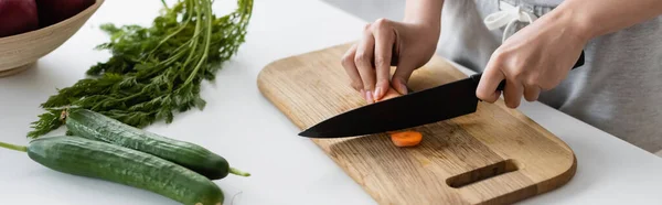 Vista recortada de la mujer cortando zanahoria cruda en la tabla de cortar cerca de pepinos frescos en la mesa, pancarta - foto de stock