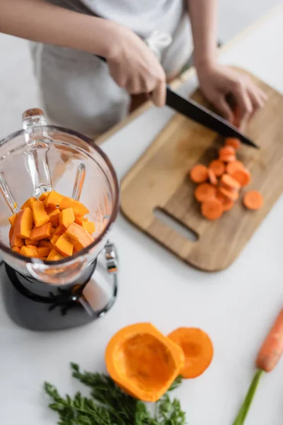 Vue partielle de femme floue coupant la carotte près du mélangeur avec la citrouille — Photo de stock