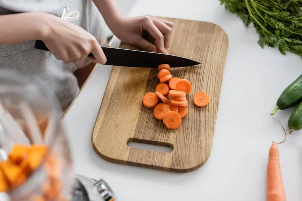 Vue partielle de la femme coupant la carotte crue près des concombres frais sur la table de cuisine — Photo de stock