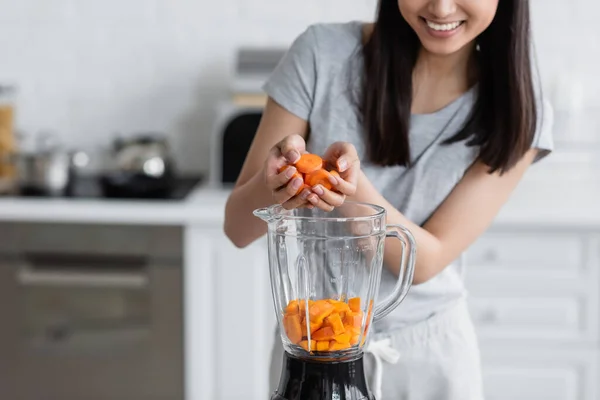 Vista recortada de la mujer sonriente añadiendo zanahoria en rodajas en la licuadora con calabaza cortada - foto de stock