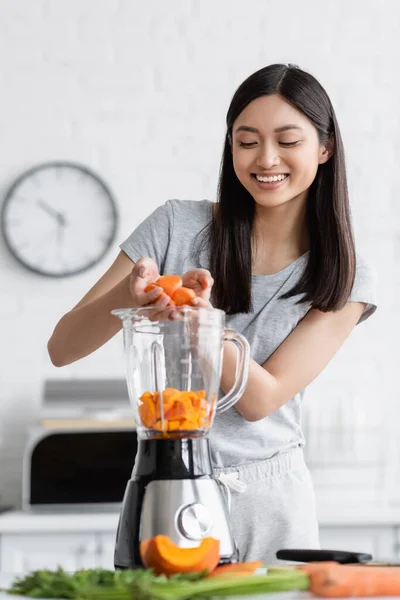 Cheerful asian woman adding sliced carrot into electric blender with cut pumpkin — Stock Photo