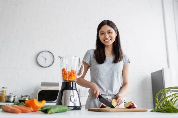 Cheerful asian woman cutting apple near electric blender and fresh vegetables in kitchen — Stock Photo