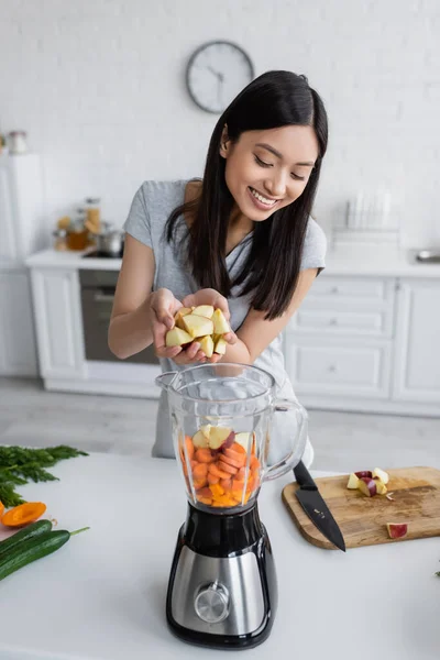 Freudige Asiatin fügt geschnittenen Apfel in elektrischen Mixer mit rohem Gemüse hinzu — Stockfoto