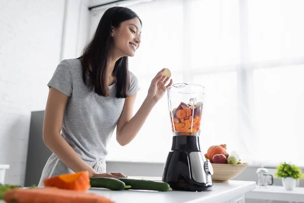 Smiling asian woman adding apple into blender with fresh vegetables in kitchen — Stock Photo