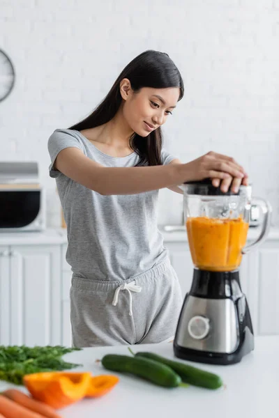 Junge Asiatin bereitet frischen Smoothie im Mixer in der Nähe verschwommenes Gemüse auf dem Tisch zu — Stockfoto