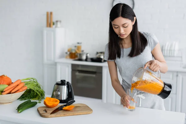 Young asian woman pouring fresh smoothie near fresh vegetables and chopping board on kitchen table — Stock Photo