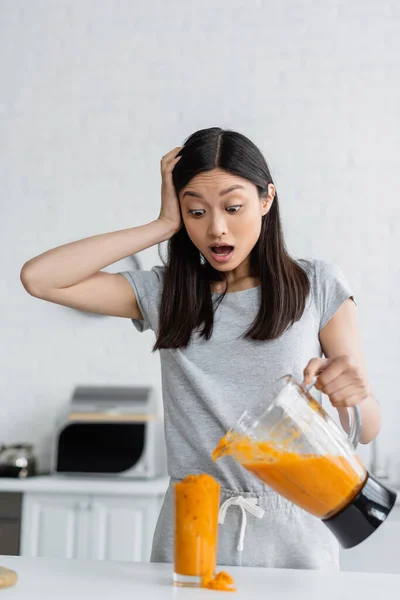 Scioccato asiatico donna toccare testa vicino traboccante vetro di frullato in cucina — Foto stock