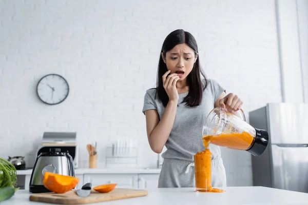 Worried asian woman pouring smoothie into overflowing glass near cut pumpkin on chopping board — Stock Photo