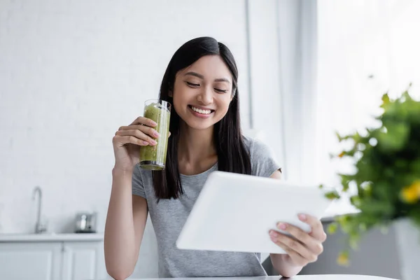 Lächelnde Asiatin blickt auf digitales Tablet, während sie ein Glas leckeren Smoothie in der Hand hält — Stockfoto