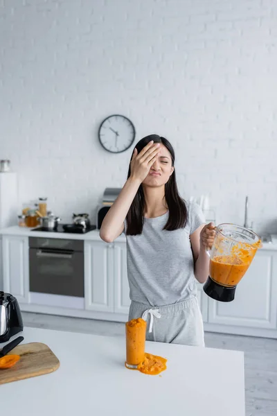 Displeased asian woman covering face with hand while holding jar of smoothie near overflowing glass — Stock Photo