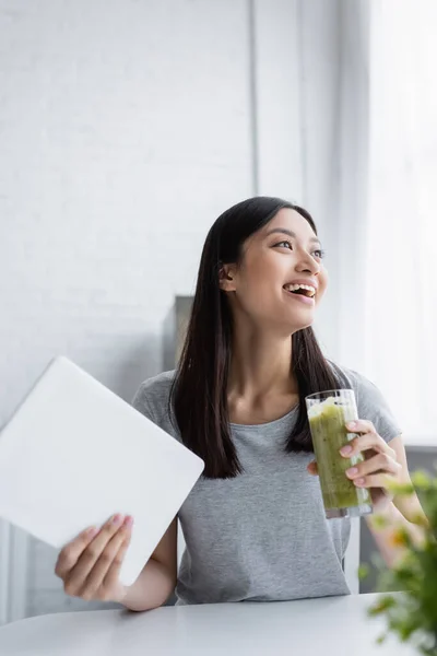 Cheerful asian woman with glass of fresh smoothie and digital tablet laughing in kitchen — Stock Photo
