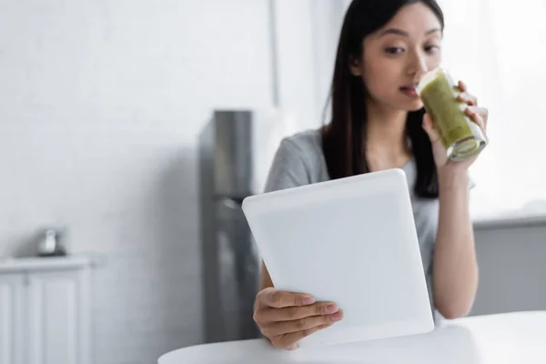 Desenfocado asiático mujer bebiendo casero smoothie mientras usando digital tablet - foto de stock