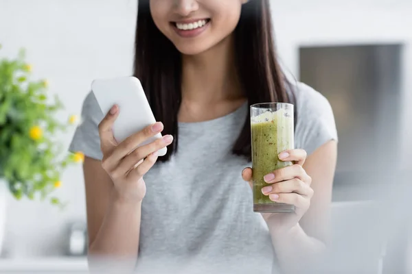 Partial view of brunette woman with glass of smoothie messaging on smartphone on blurred foreground — Stock Photo