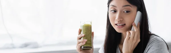 Jeune femme asiatique tenant verre de smoothie frais tout en parlant sur smartphone, bannière — Photo de stock