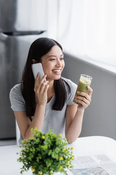 Alegre asiático mujer con vidrio de fresco smoothie hablando en móvil teléfono cerca borrosa planta - foto de stock