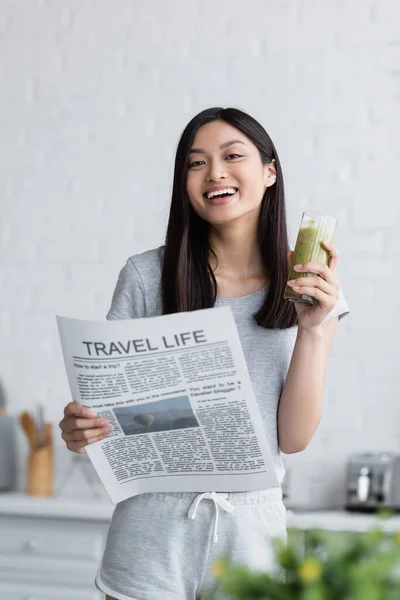 Felice donna asiatica con vita di viaggio giornale e vetro di frullato fresco guardando la fotocamera in cucina — Foto stock