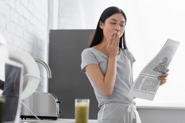 Somnolent asiatique fille bâiller tout en lisant matin journal dans cuisine — Photo de stock