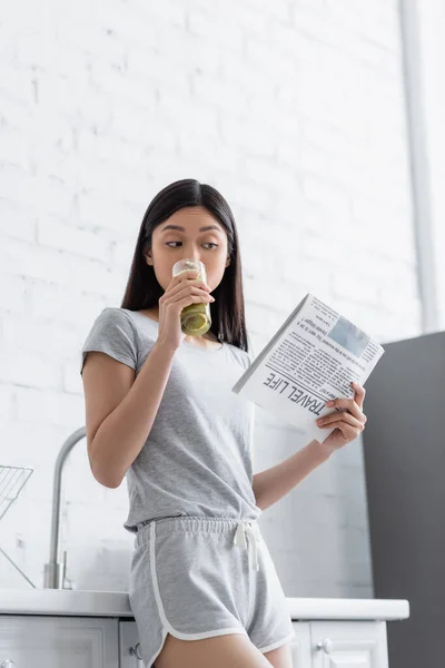 Молодая азиатская женщина пьет свежий смузи во время чтения туристической жизни газеты на кухне — стоковое фото