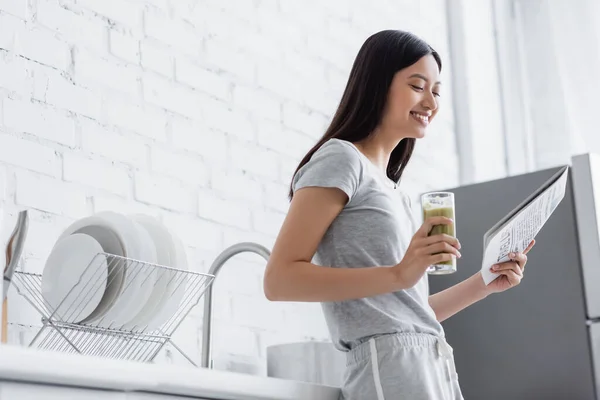 Alegre asiático mulher com vidro de fresco smoothie leitura jornal no cozinha — Fotografia de Stock