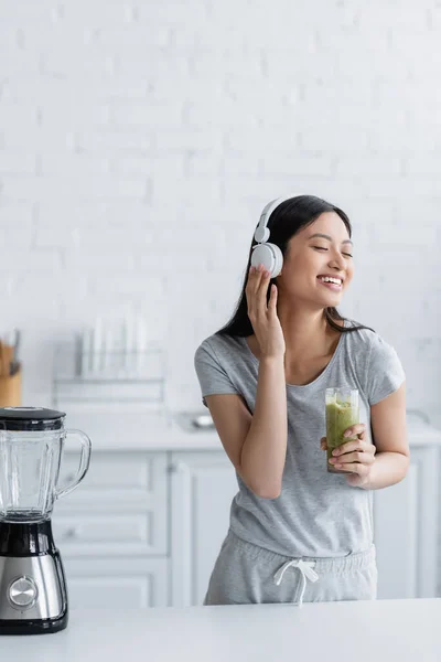 Alegre mulher asiática com vidro de smoothie ouvindo música em fones de ouvido perto de liquidificador elétrico — Fotografia de Stock