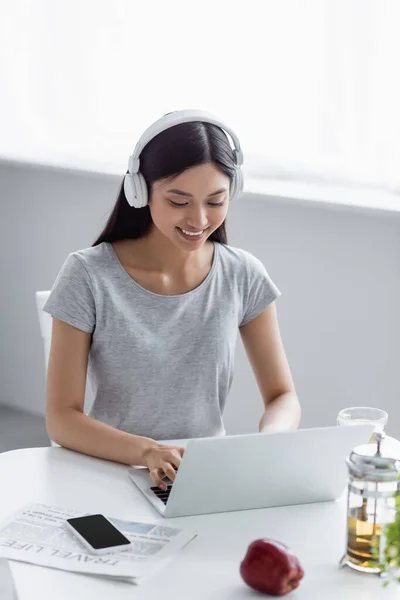 Smiling asian woman in headphones typing on laptop near teapot and smartphone on desk — Stock Photo