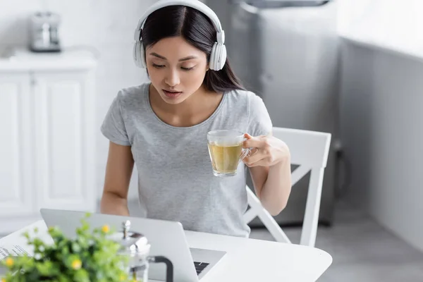 Young asian woman in headphones looking at laptop while holding cup of tea — Stock Photo
