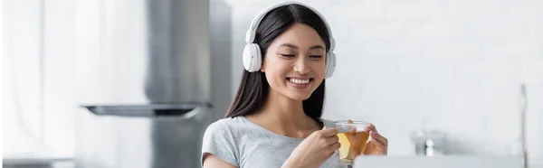 Mujer asiática feliz sosteniendo taza de té y escuchando música en los auriculares, bandera - foto de stock