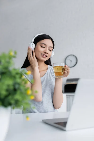 Задоволена азіатська жінка розслабляється в бездротових навушниках з чашкою чаю біля ноутбука — стокове фото