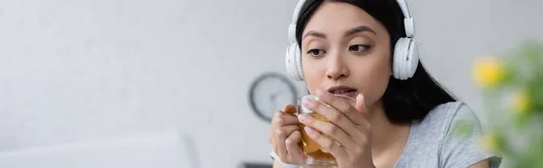 Jeune femme asiatique écoute de la musique dans les écouteurs sans fil tout en tenant une tasse de thé, bannière — Photo de stock
