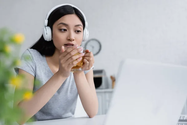 Concentré asiatique femme dans sans fil casque tenant tasse de thé près ordinateur portable — Photo de stock