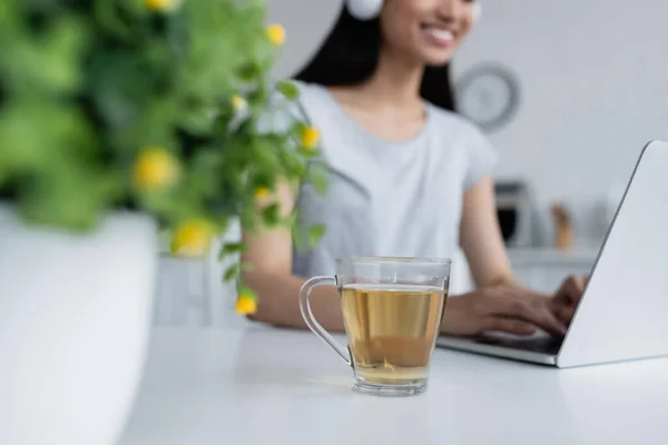 Vue recadrée de brouillé asiatique femme dactylographier sur ordinateur portable près tasse de thé — Photo de stock