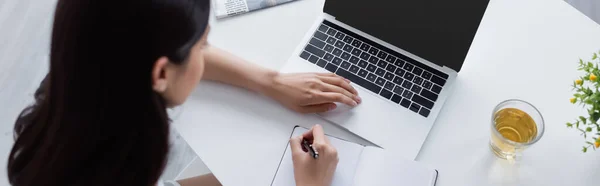 Overhead view of woman writing in notebook near laptop and cup of tea on desk, banner — Stock Photo