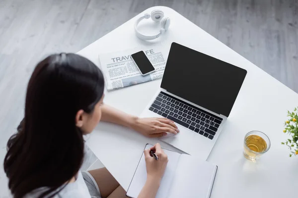 Vista aerea di donna sfocata che scrive in notebook vuoto vicino a gadget e giornali — Foto stock