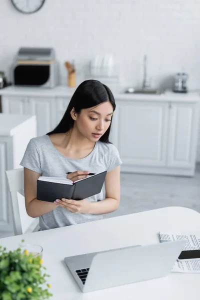 Молодая азиатка пишет в блокноте возле ноутбука на кухне — стоковое фото