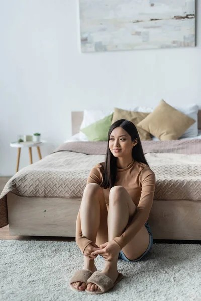 Sonriente asiático mujer sentado en piso cerca cama y mirando lejos - foto de stock