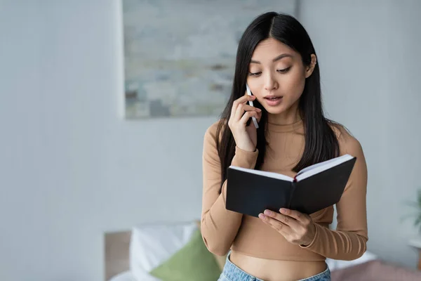 Jeune asiatique femme à la recherche dans notebook tout en parlant sur téléphone portable dans chambre — Photo de stock