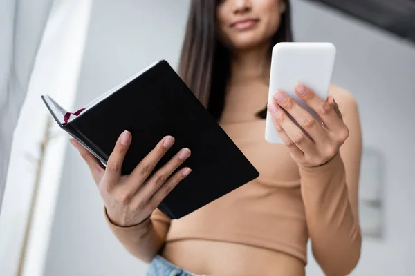 Cropped view of blurred woman holding smartphone and notebook while working at home — Stock Photo