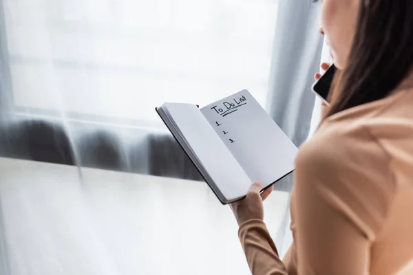 Vista cortada de mulher segurando notebook com a fazer lista de letras enquanto fala no smartphone — Fotografia de Stock