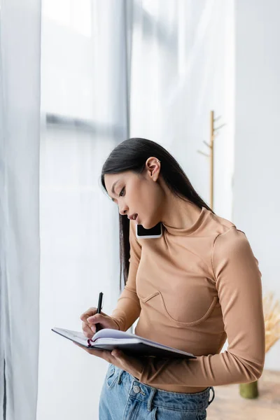 Asian woman writing in notebook while calling on smartphone at home — Stock Photo